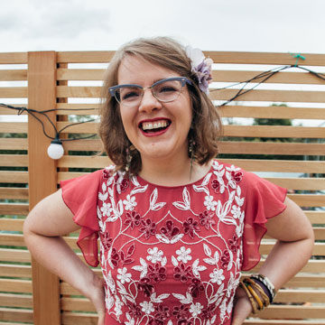Headshot of Kate McStraw in front of a fence with festoon lighting. Her hands are on her waist and she's laughter smiling. She's got a flower in her hair and cat eye framed glasses.